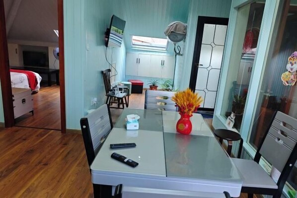 Habitación de hotel compacta con comedor, televisor montado en la pared, una mesita con un jarrón de flores y una ventana con cortinas.