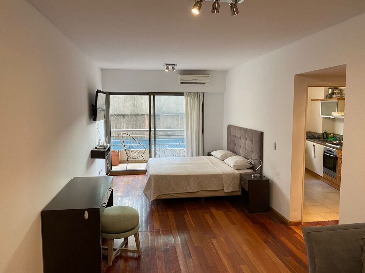 A modern hotel room with a bed, desk, and chair, a kitchenette, and a large window with a view.