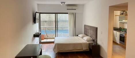 A modern hotel room with a bed, desk, and chair, a kitchenette, and a large window with a view.