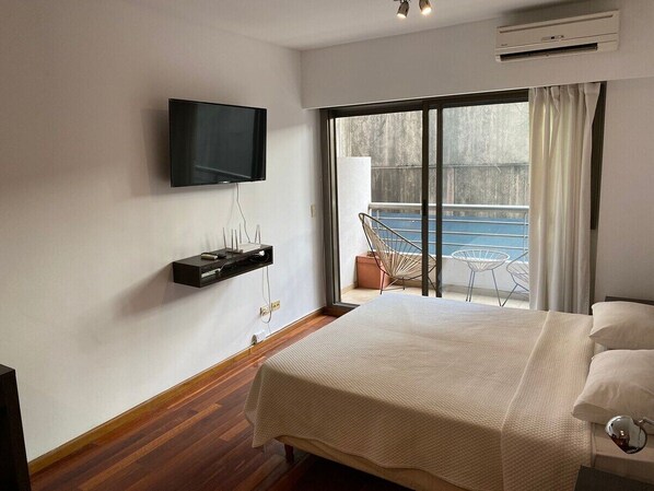 A hotel room with a bed, a TV mounted on the wall, a balcony with a chair, and a view of the sea.