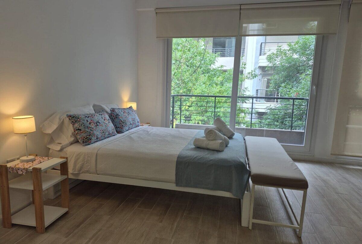 A bedroom with a bed, bedside tables, a bench, and a large window with a view of greenery and buildings.