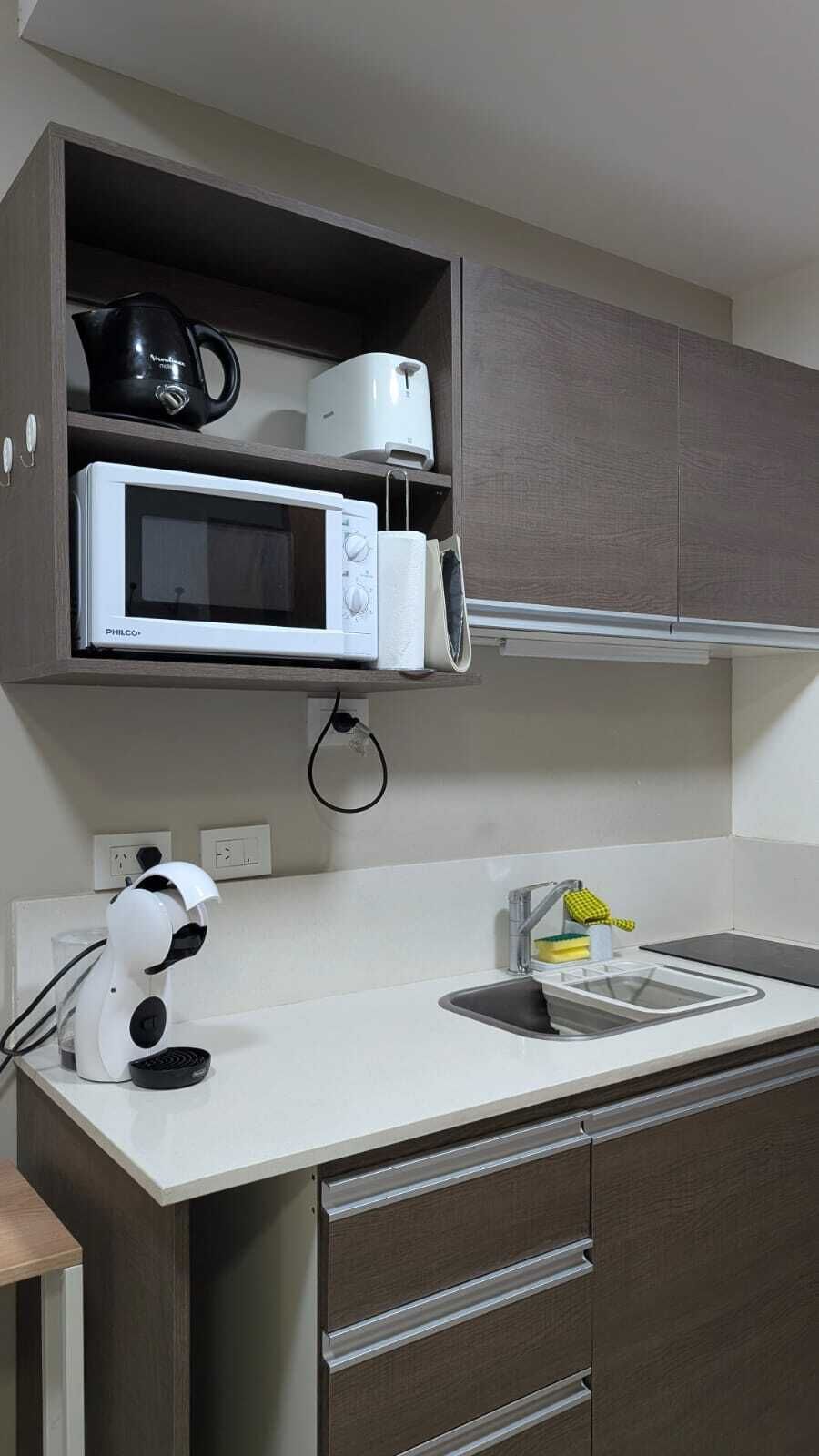 A compact kitchen with a microwave, kettle, and coffee maker on a shelf, a sink with a faucet, and a compact coffee machine.