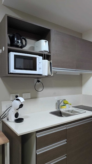 A compact kitchen with a microwave, kettle, and coffee maker on a shelf, a sink with a faucet, and a compact coffee machine.