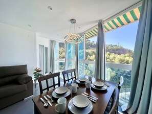A dining area with a table set for four, a sofa, and large windows offering a view of a hillside. - ANTENA POOL CITY R148 (Benidorm)