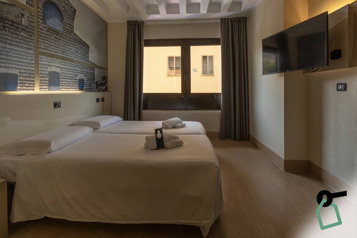 A hotel room with two beds, a large window with curtains, a flat-screen TV mounted on the wall, and a mural on the wall.