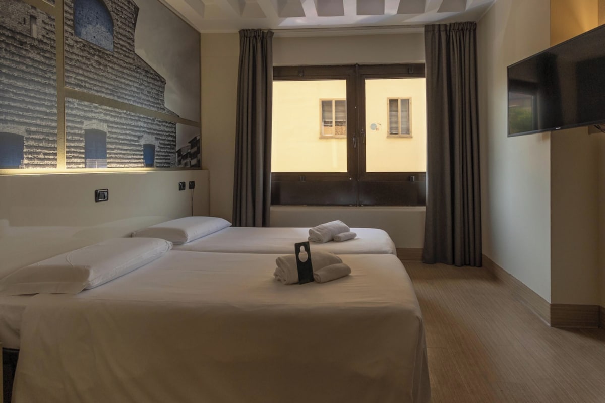Une chambre d’hôtel avec deux lits, une grande fenêtre avec des rideaux, un téléviseur à écran plat fixé au mur et une fresque murale.