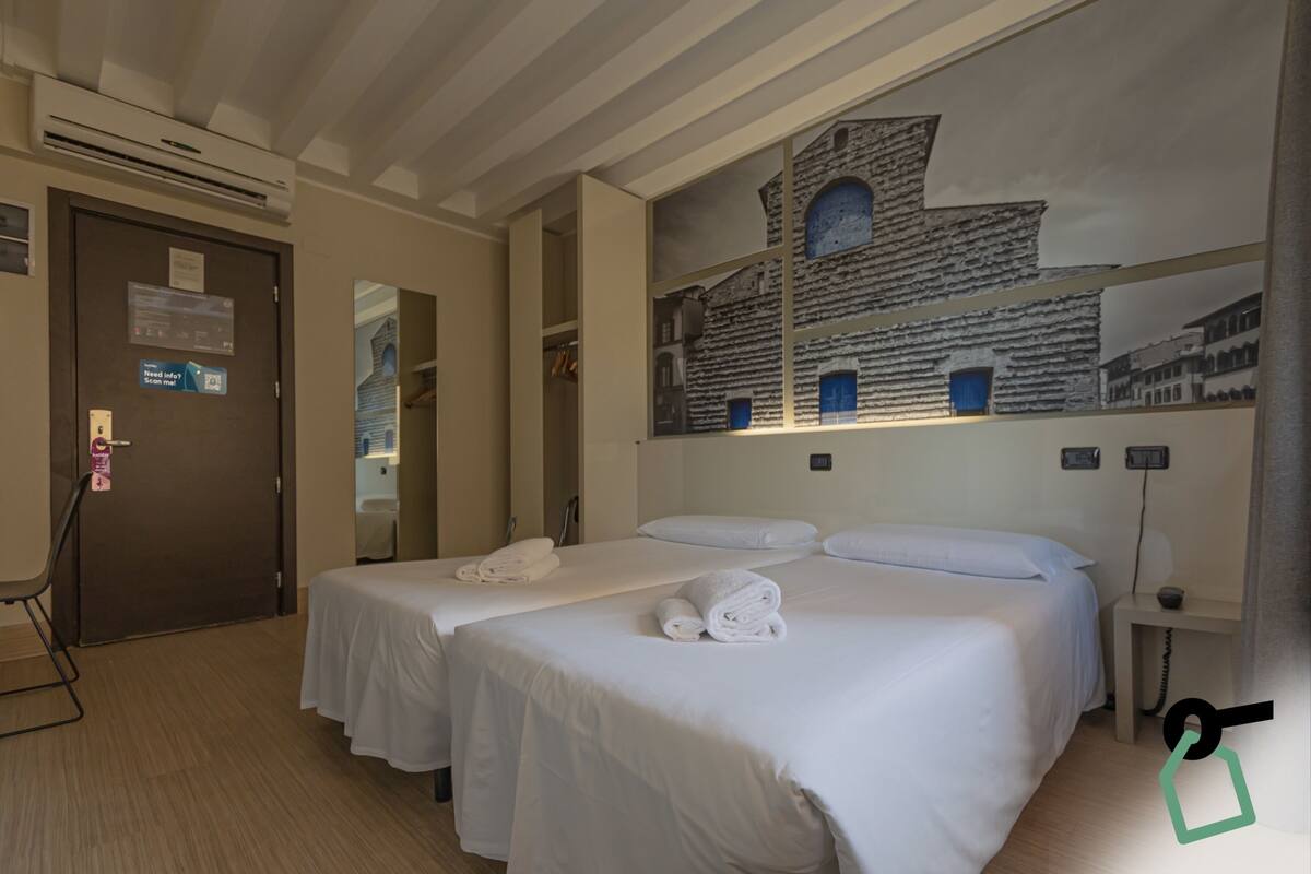 A hotel room with two beds, a large wall mural, a door with a sign, and a small table with a lamp.