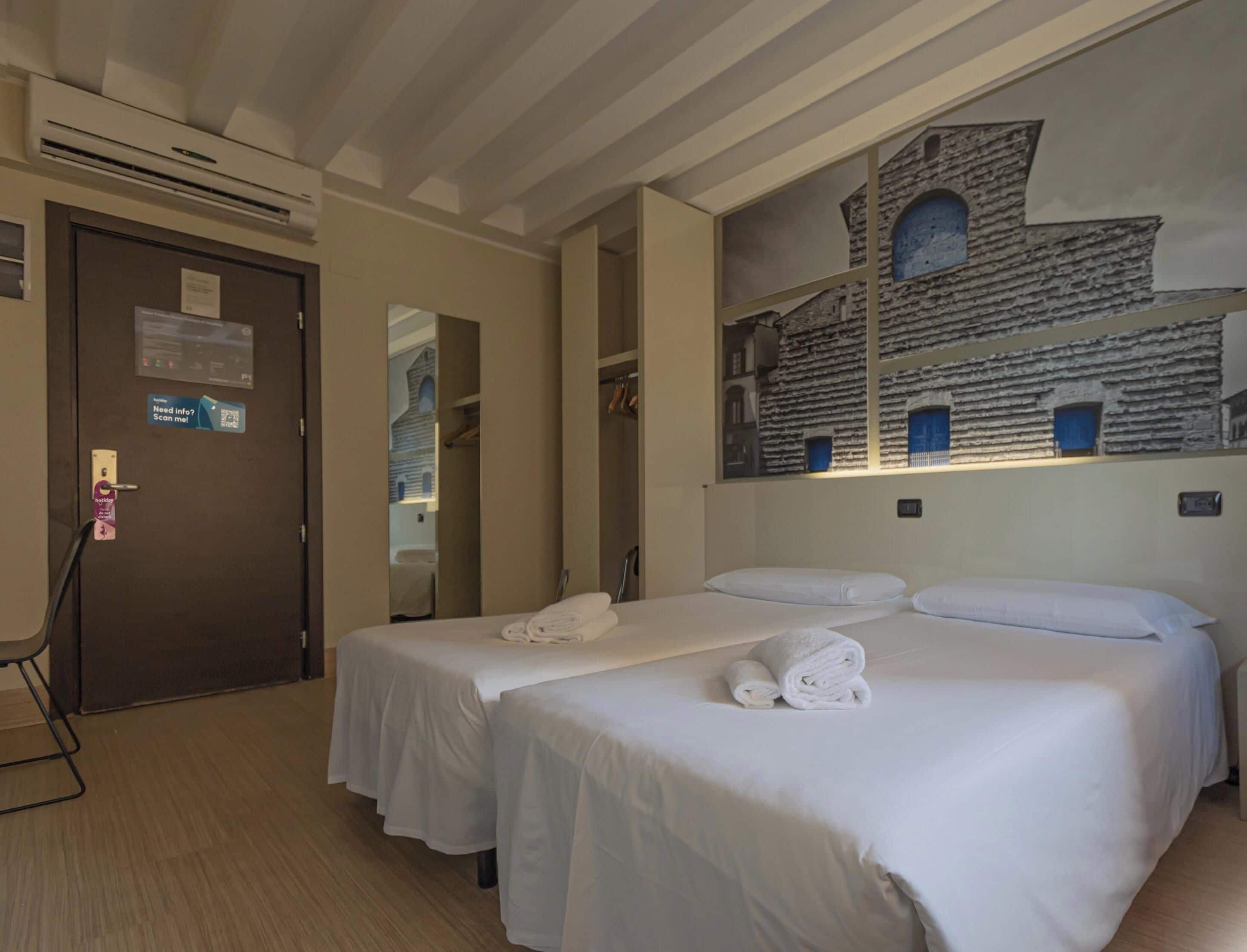 Una camera d'albergo con due letti, un grande affresco, una porta con lettore di tessere e uno specchio.