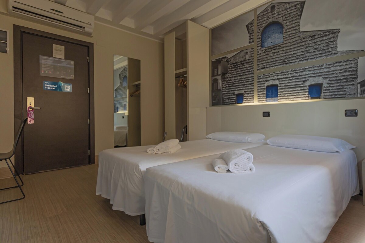 Une chambre d’hôtel avec deux lits, une grande fresque murale, une porte avec une pancarte et une petite table avec une lampe.