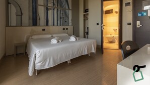A hotel room with a bed, a small bedside table, a wall-mounted artwork, a bathroom visible through an open door, and a desk with a chair. - Hotiday Firenze Guidoni (Florence)