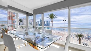 A modern dining area with a glass table and white chairs, overlooking a beach and cityscape.