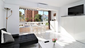 Una sala moderna con un sofá negro, una mesa de centro blanca y un televisor. Hay un balcón con vistas a edificios y palmeras.