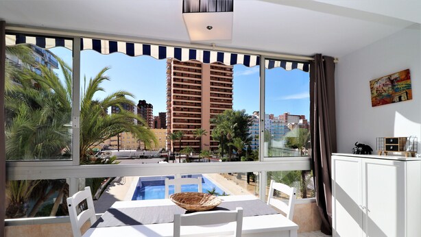 Un balcón con mesa, sillas, un cesto y vistas a una piscina y edificios altos.