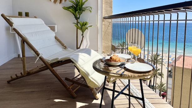 Uma varanda com uma cadeira de descanso, uma mesa pequena com prato de comida e bebidas, e vista para o mar.