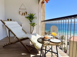 Un balcón con una silla de descanso, una mesa pequeña con comida y bebidas, y vista al mar.