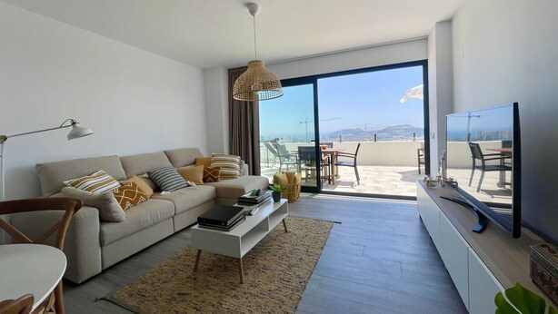 Una sala moderna con sofá, mesa de centro y televisión. Hay un balcón con vistas a la ciudad y a las montañas.