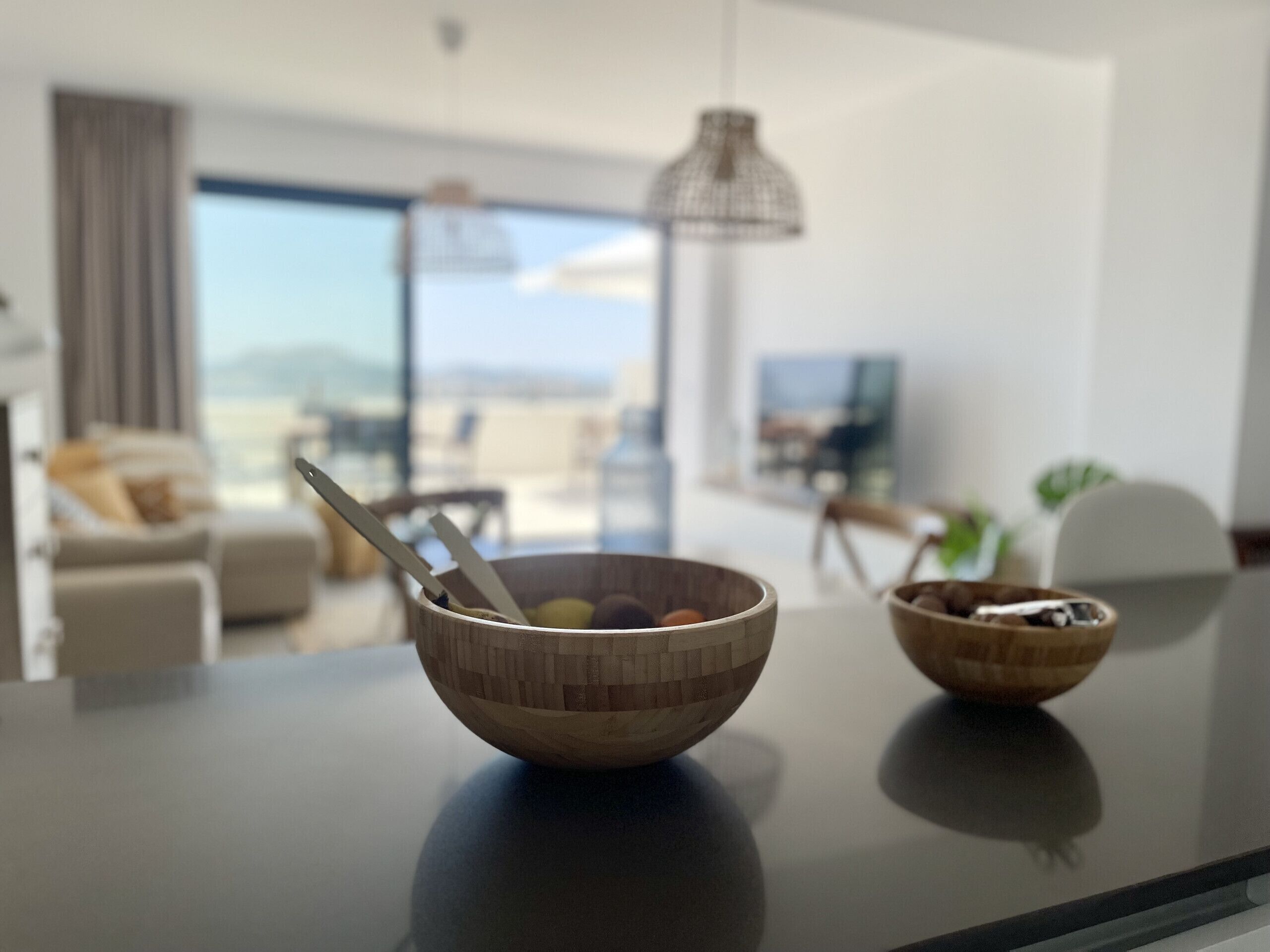 A modern living room with a large window, a dining table with a wooden bowl, and a hanging light fixture.