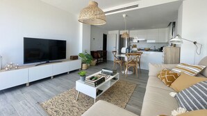 Una sala moderna con televisor de pantalla plana, una consola blanca, un sofá con cojines estampados y una zona de comedor con mesa y sillas de madera.