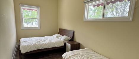A small, single-bedroom room with two beds, a wooden floor, and two windows with curtains.