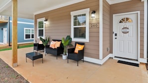 L’extérieur d’une maison avec un porche abrité, deux fauteuils en osier avec des coussins orange, une plante en pot et une porte avec une enseigne.
