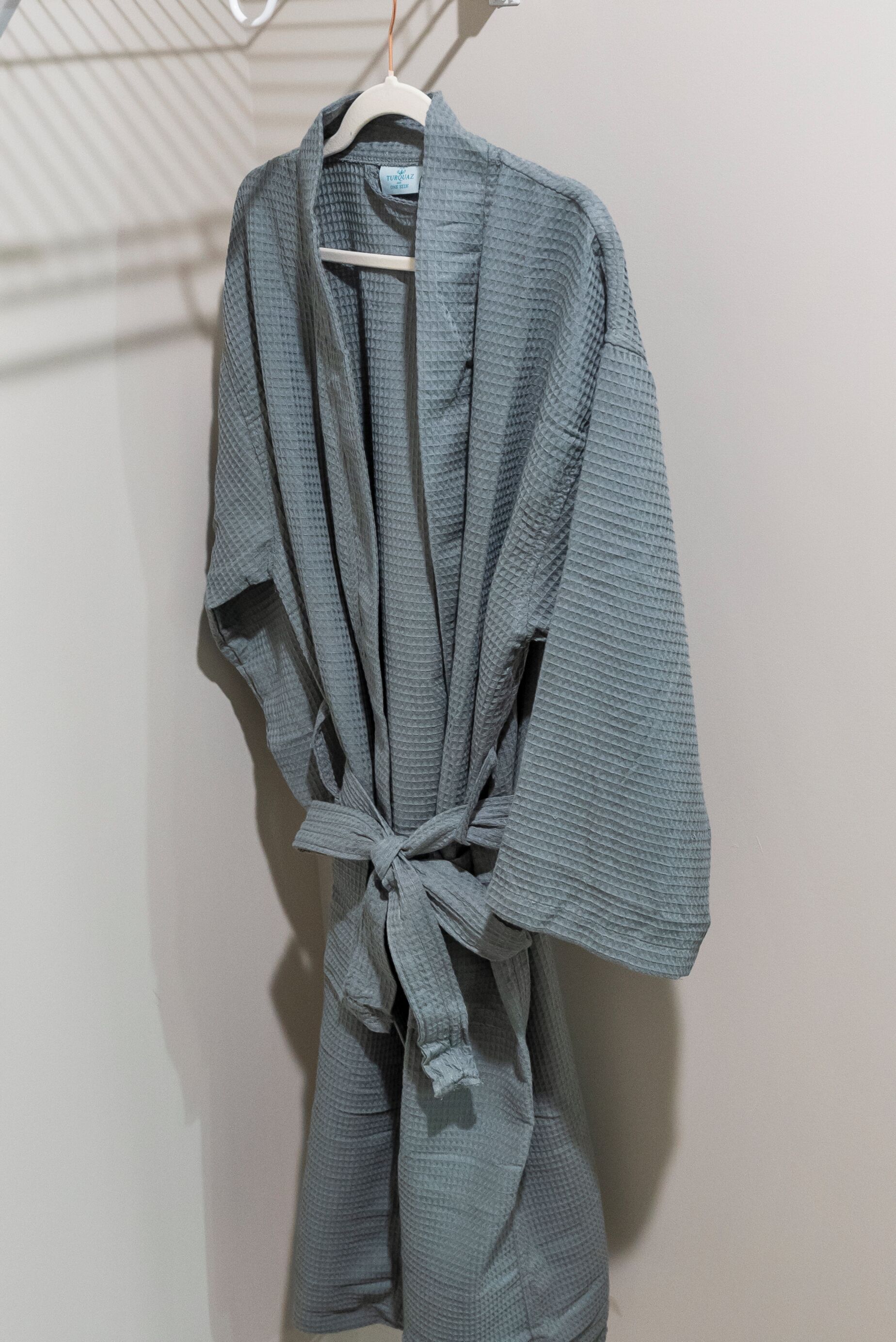 A gray robe tied at the waist, hanging on a white hanger against a plain wall.