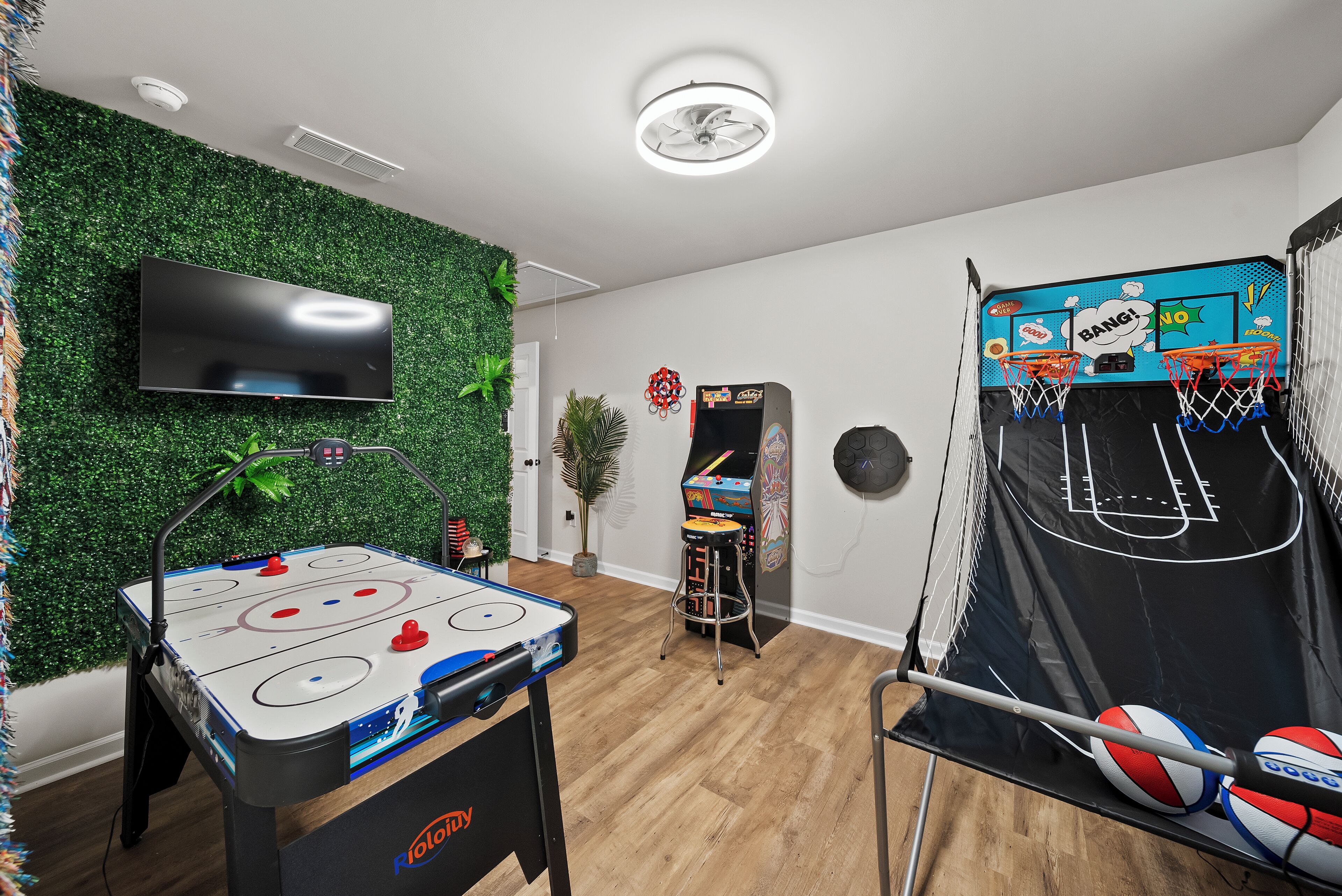 A room with a foosball table, a flat-screen TV, a basketball arcade game, and a green wall.