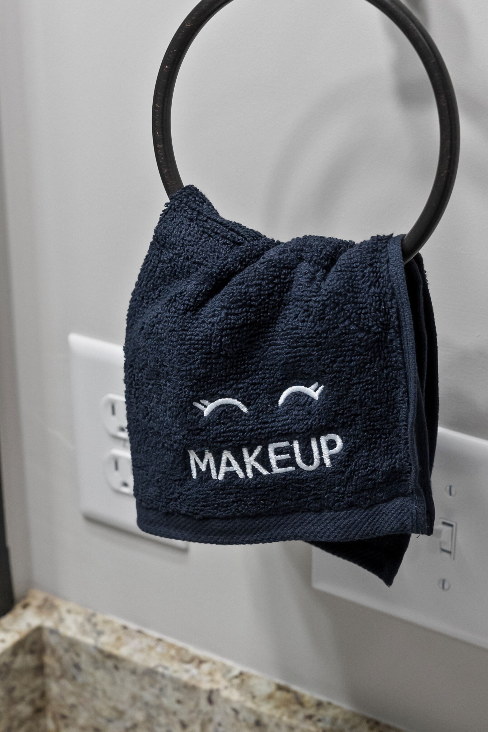 Una toalla azul oscuro con la palabra "MAKEUP" y una carita sonriente colgada de un gancho metálico.