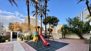 A playground with a red slide, surrounded by palm trees and a paved area with a mix of red and grey tiles. - TORRE YAGO SEAFRONT LEVANTE N028 (Benidorm)