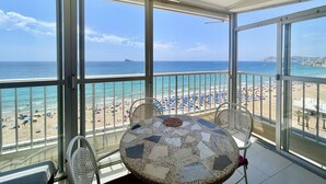Un balcon avec une table et des chaises, donnant sur une plage parsemée de parasols et où se trouvent des gens.