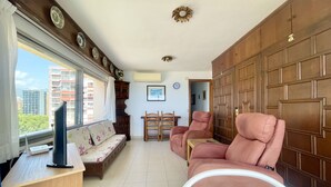 Un salon comprenant un canapé, deux fauteuils, une petite table et un téléviseur posé sur un bureau en bois. Une fenêtre offre une vue sur les bâtiments à l’extérieur.
