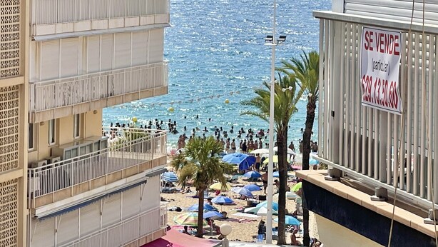 A beachfront hotel with balconies overlooking the sea, a sign indicating a sale, and beach umbrellas.