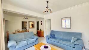 A living room with two blue sofas, a wooden coffee table, a chandelier, and a dining area with wooden chairs and a table.