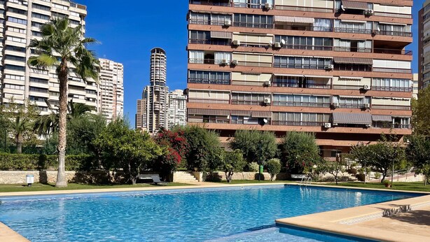 A swimming pool with a clear blue water surface, surrounded by a landscaped area with palm trees and shrubs. In the background, there are multi-story residential buildings with balconies and air conditioning units. - PRINCIPADO WIFI, AIRCO, POOL AND GARDEN N031 (Benidorm)