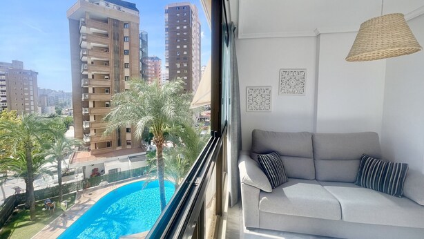 A balcony with a view of tall buildings, a swimming pool, and a grey sofa with striped pillows. - PARAISO LEVANTE BEACH STUDIO N105 (Benidorm)