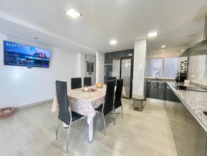 A modern kitchen with a dining area, a flat-screen TV mounted on the wall, and a trash bin.