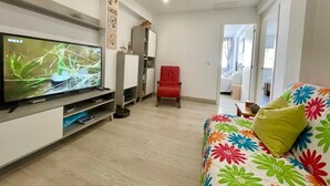 Ein Wohnzimmer mit Flachbildfernseher, einem roten Sessel, einem farbenfrohen Sofa und einem Flur, der zu einem Schlafzimmer führt.