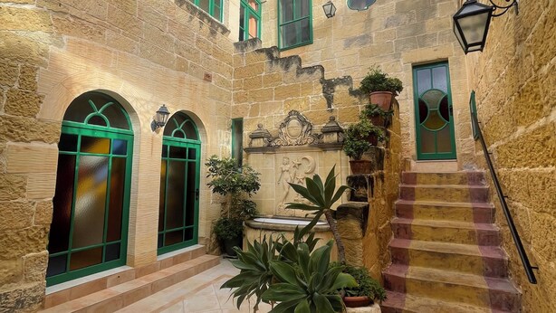 A courtyard with stone walls, green-framed windows, a fountain, and a staircase.