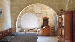 A room with a stone wall, a bed with patterned bedding, a wooden dresser, and a chair.