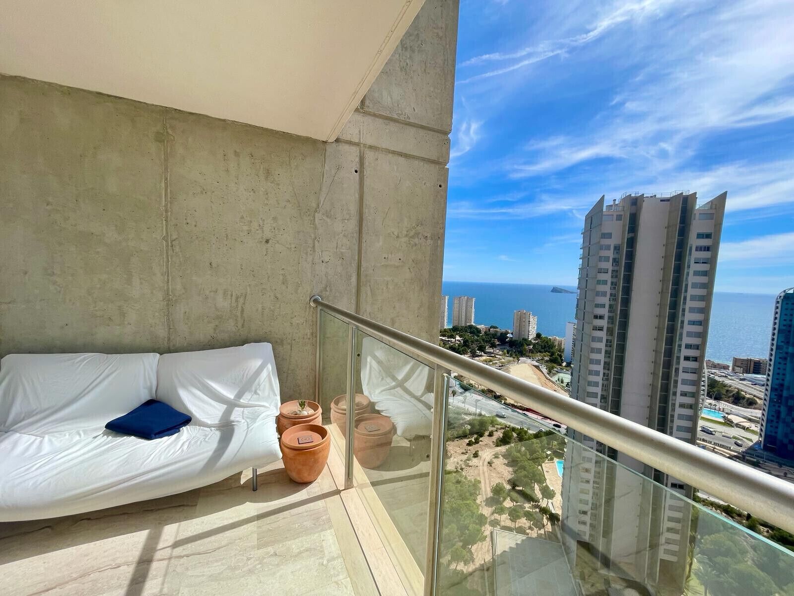 Un balcon avec un canapé blanc, des pots en terre cuite et une balustrade en verre donnant sur un paysage urbain et la mer.