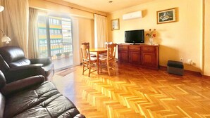 Una sala de estar con sofá de cuero, mesa de comedor de madera, sillas, un televisor en un mueble y vista a un edificio a través de una ventana.