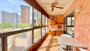 Un balcón con piso de madera, ventilador de techo, mesa de comedor con sillas y vista a edificios altos.