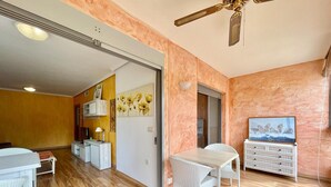 Amplia habitación con piso de madera, zona de comedor con mobiliario de mimbre blanco, ventilador de techo y televisor sobre un mueble.