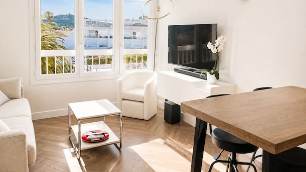 A modern living room with a TV, white furniture, a balcony with a city view, and a dining area. - Naturiste Cap d'Agde Port Soleil 146 (Agde)
