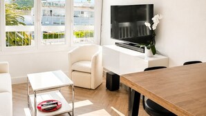 A modern living room with a TV, white furniture, and a view of buildings through large windows. - Naturiste Cap d'Agde Port Soleil 146 (Agde)