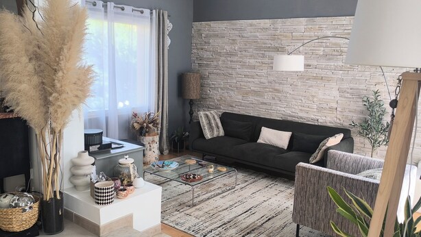 A modern living room with a stone wall, a black sofa, a glass coffee table, and decorative items.