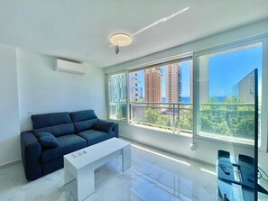 A modern living room with a dark blue sofa, a white coffee table, and a large window offering a view of tall buildings and greenery. - MAGNIFICENT MEDITERRANEO STUDIO (N113) (Benidorm)