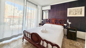 Ein Hotelzimmer mit einem großen Bett, einem Holz-Kopfteil, einem Nachttisch mit Lampe, einem Fenster mit Vorhängen und einer Wand-Klimaanlage.