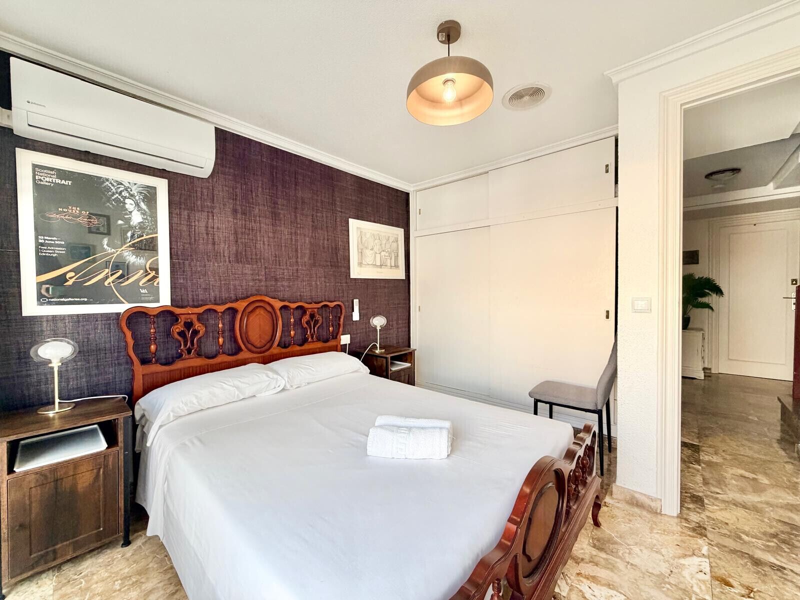 A hotel room with a large bed, wooden headboard, a nightstand, a bedside table, a wall-mounted lamp, and a framed picture.
