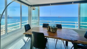 Comedor con mesa y sillas de madera, ventanas amplias con vistas al océano y una lámpara de techo moderna.