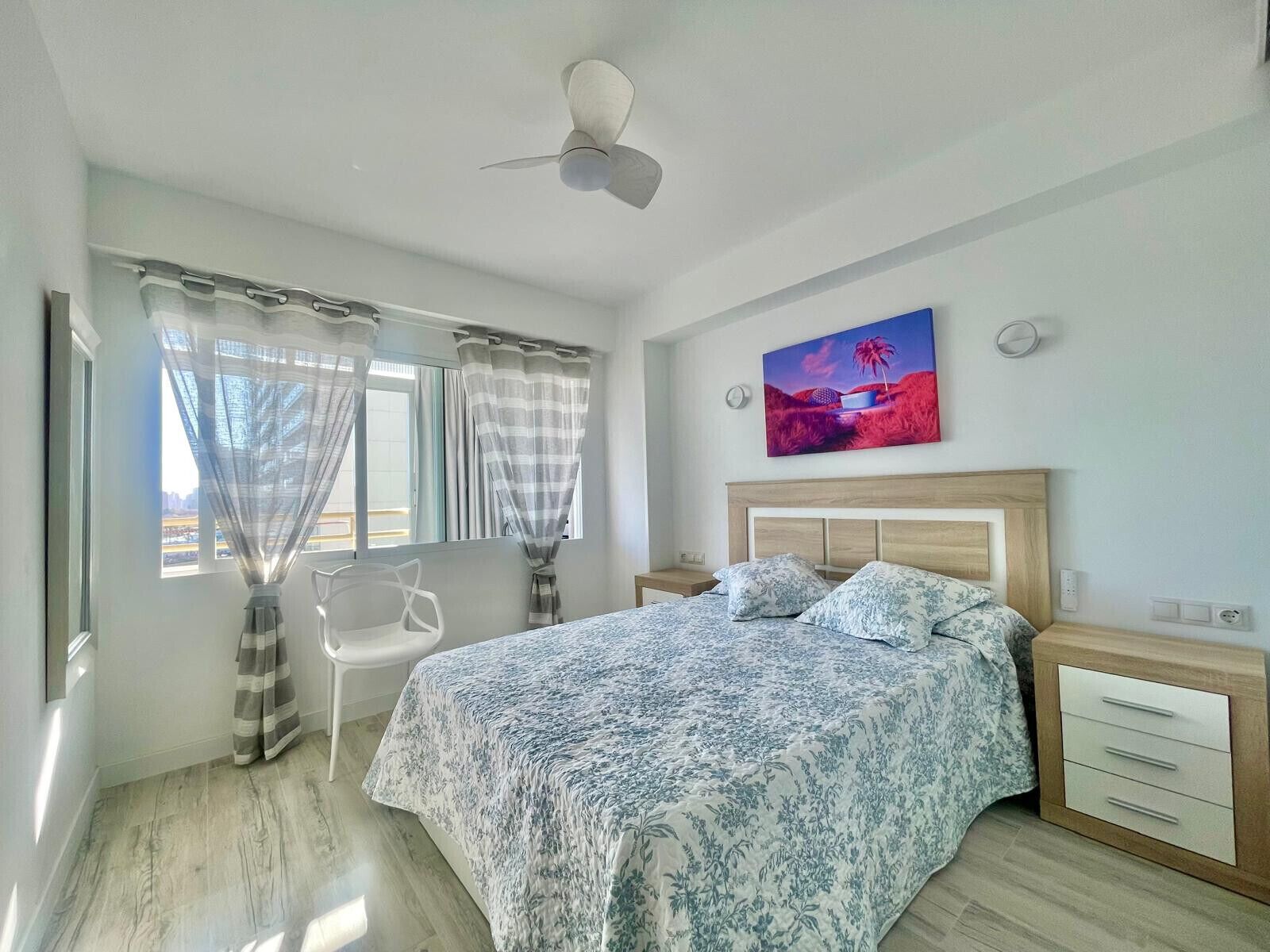 A bedroom with a bed, a chair, a nightstand, a painting, and a ceiling fan.